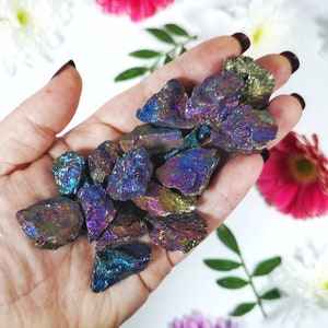 May include: A collection of iridescent pyrite stones, showcasing a range of colours such as blues, purples, and golds. The raw stones are held against a backdrop of white and colourful flowers, emphasising their natural beauty.