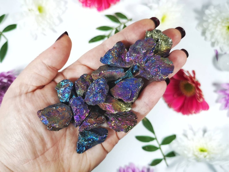 May include: A collection of iridescent pyrite stones, showcasing a range of colours such as blues, purples, and golds. The raw stones are held against a backdrop of white and colourful flowers, emphasising their natural beauty.