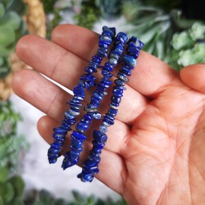 May include: Four strands of lapis lazuli chip bead bracelets. The beads are a deep blue color with flecks of lighter blue and gray. The bracelets are held in a hand, with a soft focus background of greenery.