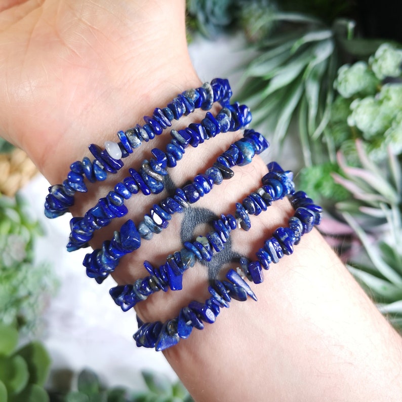 May include: Several blue lapis lazuli chip bead bracelets are stacked on a wrist. The bracelets are made of small, irregular pieces of the deep blue stone. The bracelets are strung together, creating a layered look. The background features green plants.