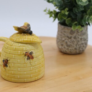 Puede incluir: Un tarro de miel de cerámica amarilla con forma de colmena, con una tapa coronada por una figura de abeja. El tarro tiene un asa y está decorado con dos abejas pintadas. El tarro está sobre una superficie de madera.