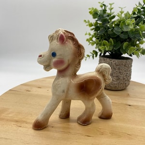 May include: A ceramic figurine of a white horse with brown markings. The horse has blue eyes and pink cheeks. It is standing on a wooden surface.