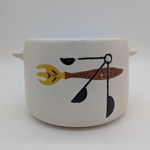 May include: Cream-colored ceramic bowl with two small handles. The bowl features a whimsical design of a fork, spoon, and other kitchen utensils in yellow, brown, and black. The bowl is suitable for serving food.