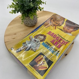 May include: A vintage Raggedy Ann Push Button Marionette toy in its original packaging. The box is yellow with images of children and the Raggedy Ann doll. The packaging includes the text "Push Button Marionette" and "US Patent No. 3178852".