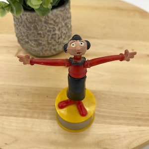 May include: A vintage plastic puppet of a woman with black hair, wearing a red shirt, black skirt, and red shoes. The puppet is standing on a yellow base with a gray ring.