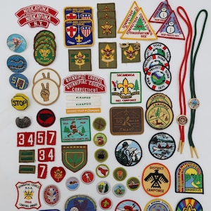 May include: A collection of vintage Boy Scout patches and pins, featuring various designs and text. Includes patches with text like "Niskayuna N.Y.", "Camp Boyhaven", and "Deep Freeze". Also includes a bolo tie with a circular emblem.