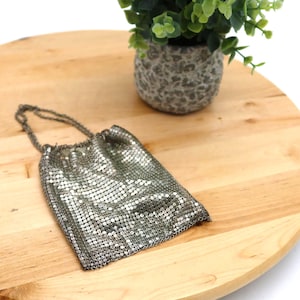 May include: A silver mesh evening bag with a drawstring closure rests on a light wood surface. A small potted plant with green foliage sits in the background. The bag has a chain handle.