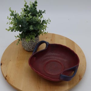 May include: A red ceramic serving bowl with two blue handles. The bowl is sitting on a wooden surface.