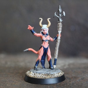 May include: A painted miniature figurine of a female demon with white hair, red skin, and a blue and black outfit. She is holding a bone staff with a silver trident at the top. The figurine is standing on a round base.