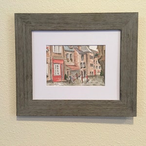 May include: A watercolor painting of a European street scene with buildings, a cafe, and people walking by. The buildings are painted in shades of brown and red, and the cafe has a red awning. The people are painted in a simple style, and they are wearing casual clothing.