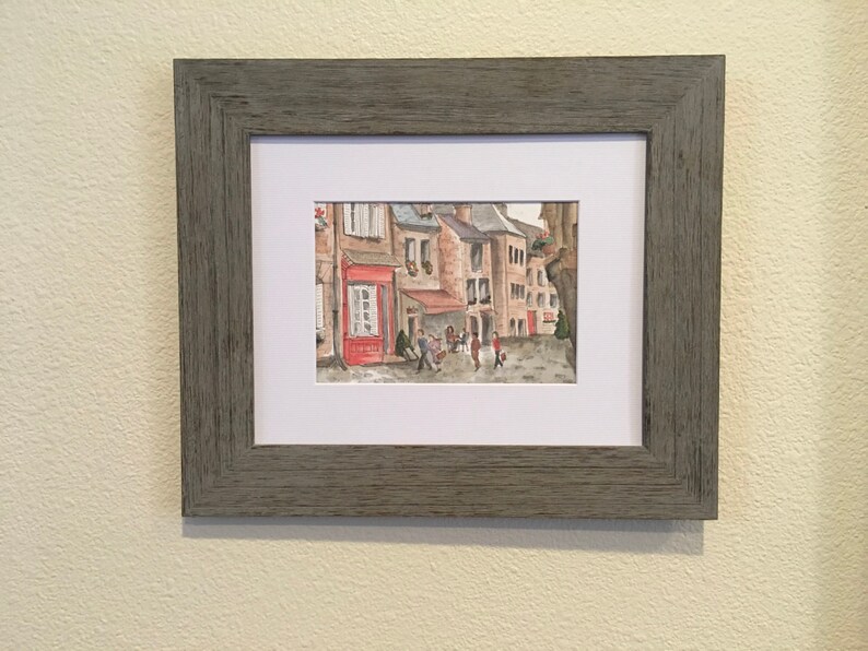 May include: A watercolor painting of a European street scene with buildings, a cafe, and people walking by. The buildings are painted in shades of brown and red, and the cafe has a red awning. The people are painted in a simple style, and they are wearing casual clothing.