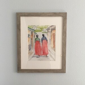 May include: A watercolor painting of two people in red dresses walking down a narrow street. The painting is framed in a light gray frame.