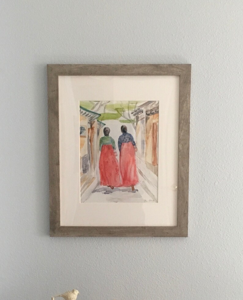 May include: A watercolor painting of two people in red dresses walking down a narrow street. The painting is framed in a light gray frame.