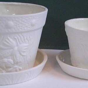 May include: Two off-white ceramic flower pots with matching saucers. The pots have a textured floral design and a decorative rim. The saucers have a matching textured pattern. The pots are different sizes.