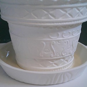 Puede incluir: Una maceta de cerámica blanca con un platillo a juego. La maceta tiene un patrón en relieve decorativo de diamantes y diseños florales. El platillo es redondo y tiene un patrón en relieve similar. La maceta se asienta sobre el platillo.