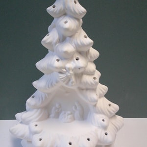 Könnte beinhalten: Ein weißer Weihnachtsbaum aus Keramik mit detailliertem Design. Der Baum hat mehrere Astschichten mit kleinen Löchern und einen Stern oben. Eine Krippenszene ist am Fuß des Baumes zu sehen. Der Baum ist etwa 25 cm hoch.