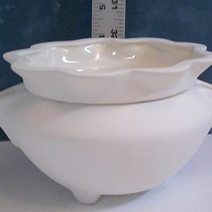 Può includere: Una fioriera in ceramica bianca con bordo smerlato, con un piattino abbinato sopra. La fioriera ha una base arrotondata e tre piedini. Il piattino misura circa 13 cm di diametro.