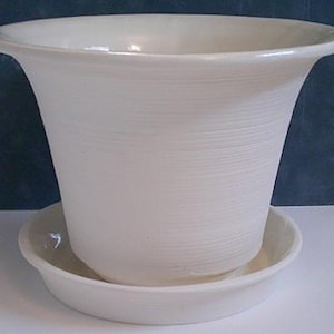 Puede incluir: Una maceta de cerámica blanca con un platillo a juego. La maceta tiene una parte superior acampanada y un patrón de rayas horizontales texturizadas. El platillo redondo se encuentra debajo, creando un conjunto para plantas de interior o exterior.