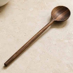 Può includere: Un cucchiaio di legno a manico lungo realizzato in legno di noce marrone scuro con una venatura naturale visibile. Il cucchiaio presenta una coppa arrotondata e liscia e un manico dritto e sottile, che mette in risalto i toni ricchi del legno.