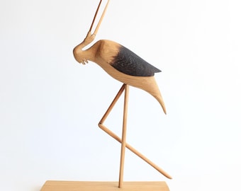 May include: Wooden sculpture of a bird with a long neck and black and brown feathers standing on a wooden base.