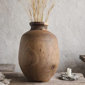 May include: A large, wooden vase with a wide base and narrow neck, filled with dried wheat stalks. The vase sits on a rustic wooden table, with a neutral-toned background. A candle and small stool are also visible.