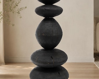 May include: A vertical sculpture composed of five stacked, rounded, black wooden forms. The shapes vary in size, with a large central oval piece flanked by flatter, disc-like elements at the top and bottom, all featuring a textured, dark finish.