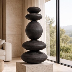 May include: A modern sculpture featuring a stack of five dark, oval-shaped stones atop a square, light-colored pedestal. The stones vary in size and are arranged to create a balanced, artistic display. The sculpture is a decorative element for interior spaces.