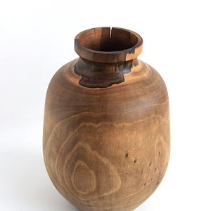 May include: A handcrafted wooden vase with a rounded body and a narrow neck. The vase is made of light brown wood with darker brown accents and visible wood grain. The vase is approximately 10 inches tall and 8 inches wide, a decorative item.
