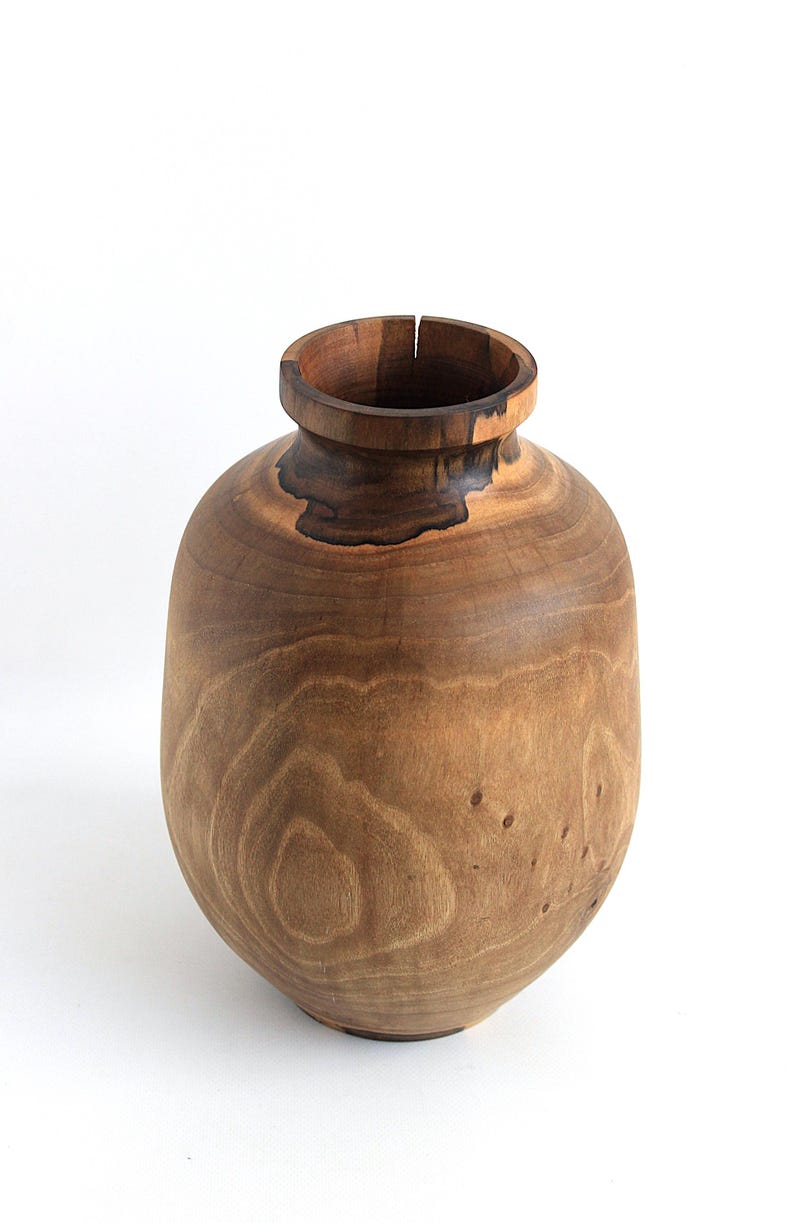 May include: A handcrafted wooden vase with a rounded body and a narrow neck. The vase is made of light brown wood with darker brown accents and visible wood grain. The vase is approximately 10 inches tall and 8 inches wide, a decorative item.