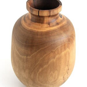 May include: A handcrafted wooden vase with a rounded body and a narrow neck. The vase is made of light brown wood with visible grain patterns. The interior of the vase is a darker brown. The vase is a decorative item.