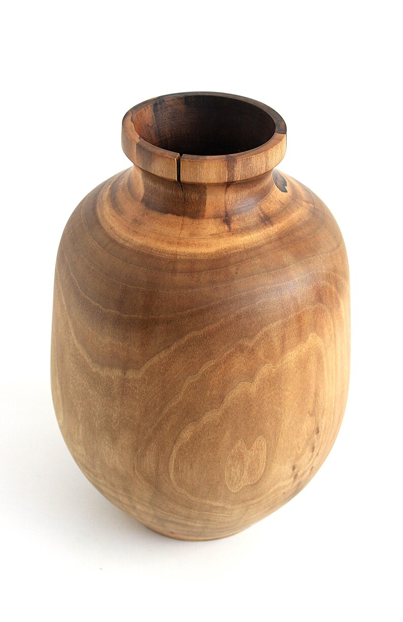 May include: A handcrafted wooden vase with a rounded body and a narrow neck. The vase is made of light brown wood with visible grain patterns. The interior of the vase is a darker brown. The vase is a decorative item.