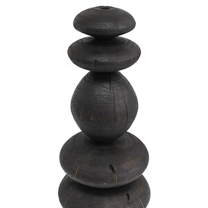 May include: A dark, stacked wooden sculpture with a natural, weathered appearance. The sculpture is composed of five rounded, disc-shaped pieces and one oval-shaped piece, all in varying sizes. The wood grain is visible, and the overall effect is rustic and organic.