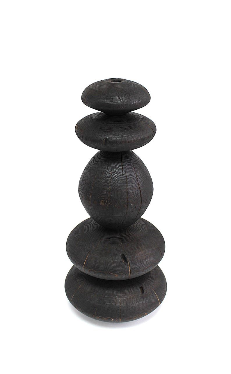 May include: A dark, stacked wooden sculpture with a natural, weathered appearance. The sculpture is composed of five rounded, disc-shaped pieces and one oval-shaped piece, all in varying sizes. The wood grain is visible, and the overall effect is rustic and organic.