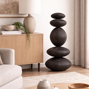 May include: A modern living room scene featuring a stacked stone sculpture, a beige lamp, and a wooden cabinet. The sculpture is composed of five dark, oval-shaped stones. A beige sofa and a round wooden coffee table complete the decor.