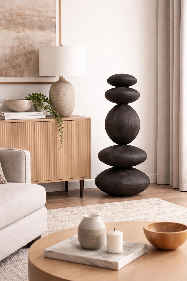 May include: A modern living room scene featuring a stacked stone sculpture, a beige lamp, and a wooden cabinet. The sculpture is composed of five dark, oval-shaped stones. A beige sofa and a round wooden coffee table complete the decor.