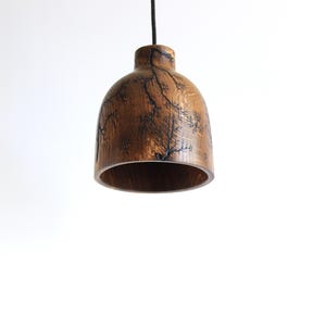 Peut inclure: Une suspension en forme de cloche fabriqu&eacute;e en bois brun poli. La surface ext&eacute;rieure pr&eacute;sente des motifs fractals sombres et complexes cr&eacute;&eacute;s par pyrogravure haute tension, suspendue par un fin cordon noir.