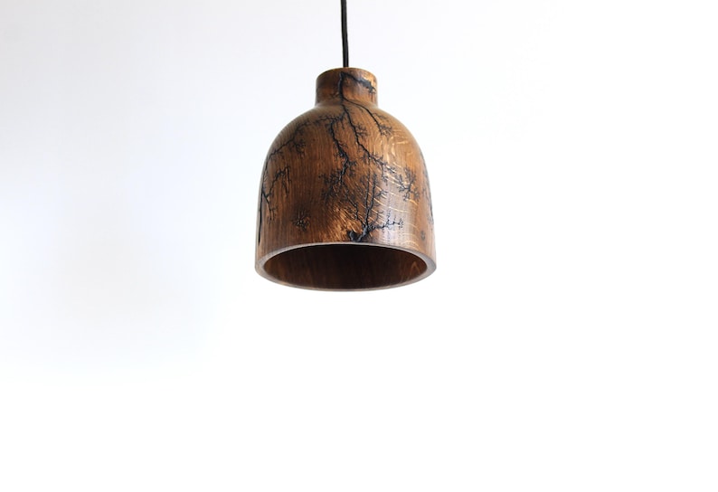 Peut inclure: Une suspension en forme de cloche fabriqu&eacute;e en bois brun poli. La surface ext&eacute;rieure pr&eacute;sente des motifs fractals sombres et complexes cr&eacute;&eacute;s par pyrogravure haute tension, suspendue par un fin cordon noir.
