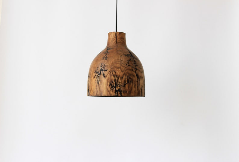 Peut inclure: Une suspension en forme de cloche fabriqu&eacute;e en bois naturel, pr&eacute;sentant un grain apparent et des marques de br&ucirc;lure fractales sombres et ramifi&eacute;es sur la surface, suspendue par un fin cordon noir sur un fond blanc uni.
