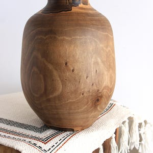 May include: A wooden vase with a wide, rounded body and a narrow neck. The vase is made of light brown wood with visible grain and knots. It sits on a white and cream-colored fringed textile.