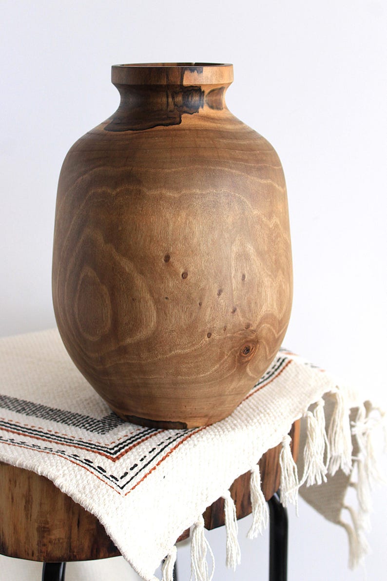 May include: A wooden vase with a wide, rounded body and a narrow neck. The vase is made of light brown wood with visible grain and knots. It sits on a white and cream-colored fringed textile.