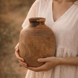 May include: A handcrafted wooden vase with a rounded body and a dark brown rim. The vase is made of light brown wood and has a smooth, natural finish. The vase is held in front of a blurred background.