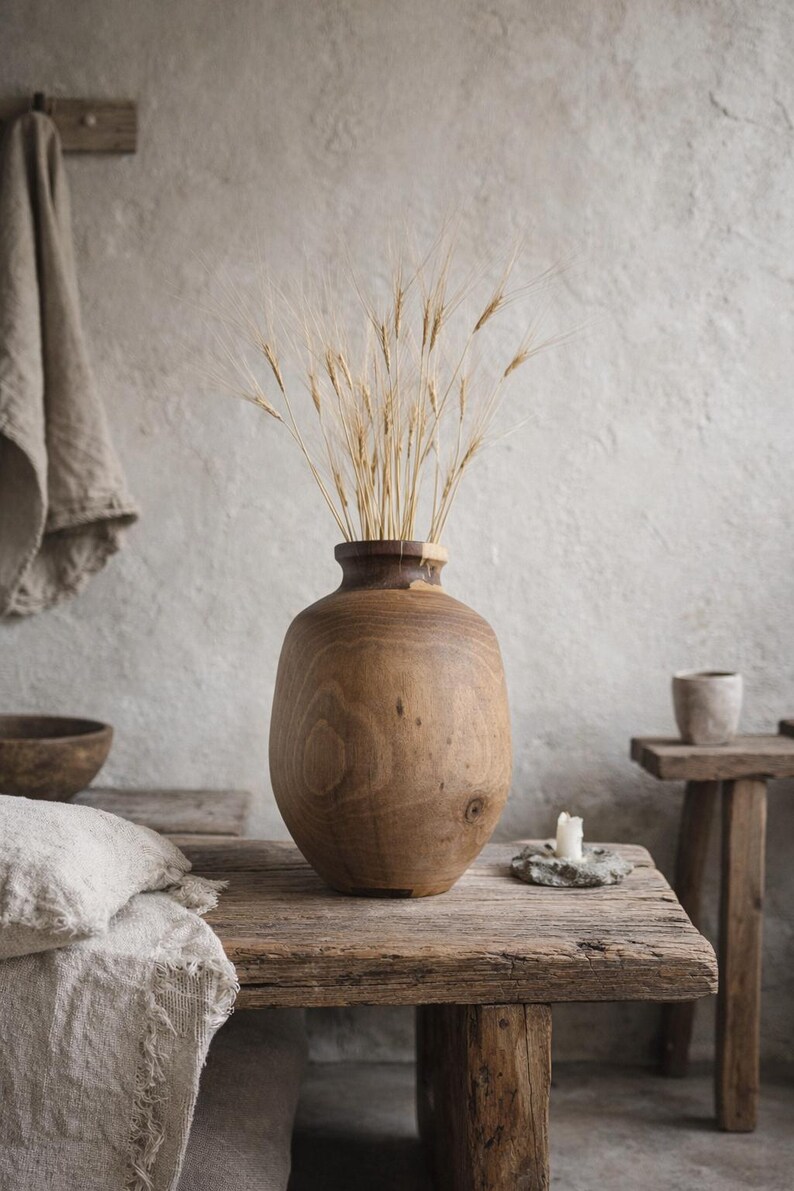 May include: A large, wooden vase with a wide base and narrow neck, filled with dried wheat stalks. The vase sits on a rustic wooden table, with a neutral-toned background. A candle and small stool are also visible.