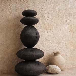 May include: A stack of dark, oval-shaped wooden decorative stones, balanced vertically. A small, beige ceramic vase and a light-colored stone sit beside the stack. The background is a textured, light brown wall.
