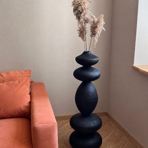 May include: A tall, black decorative vase with a stacked stone design. The vase is filled with dried, beige-colored ornamental grasses. The vase is placed next to an orange sofa in a room with wood flooring.