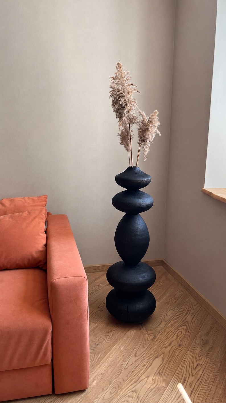 May include: A tall, black decorative vase with a stacked stone design. The vase is filled with dried, beige-colored ornamental grasses. The vase is placed next to an orange sofa in a room with wood flooring.