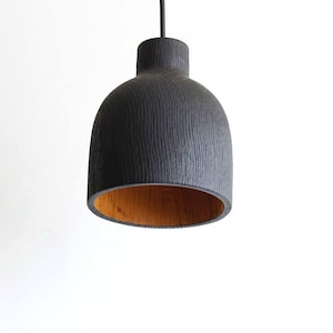 Peut inclure: Une suspension en forme de cloche avec un ext&eacute;rieur noir mat pr&eacute;sentant un motif de grain textur&eacute; vertical et une finition int&eacute;rieure en bois brun clair naturel contrastant, suspendue par un fin cordon noir sur fond blanc.