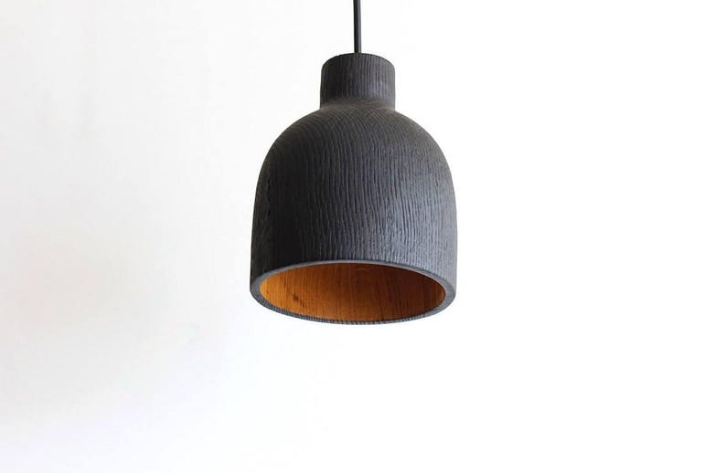 Peut inclure: Une suspension en forme de cloche avec un ext&eacute;rieur noir mat pr&eacute;sentant un motif de grain textur&eacute; vertical et une finition int&eacute;rieure en bois brun clair naturel contrastant, suspendue par un fin cordon noir sur fond blanc.