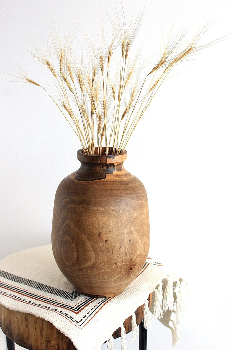 May include: A wooden vase with a wide, rounded body and a narrow neck, filled with dried wheat stalks. The vase is a warm brown color, and sits on a small wooden table covered with a white and black striped cloth.