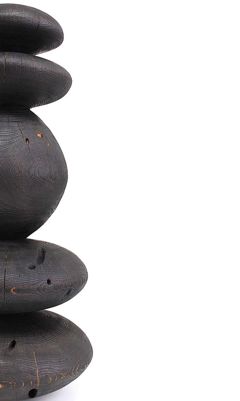 May include: A tall, dark wooden sculpture with a stacked, organic form. The sculpture is made of dark, possibly charred, wood, with a visible grain and natural imperfections. The design features rounded, stacked elements, creating a unique, abstract shape.