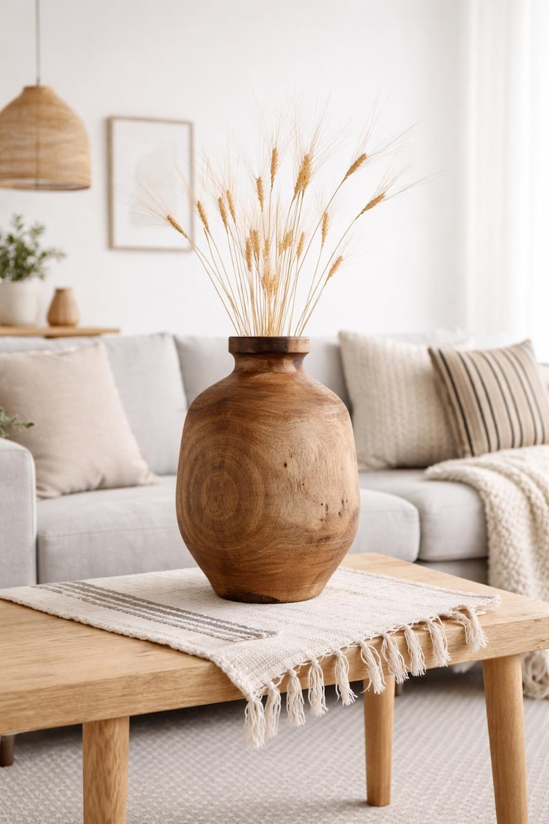 May include: A large, wooden vase with a wide, rounded body and a narrow neck. The vase is filled with dried wheat stalks. The vase sits on a light-colored wooden table with a decorative runner.